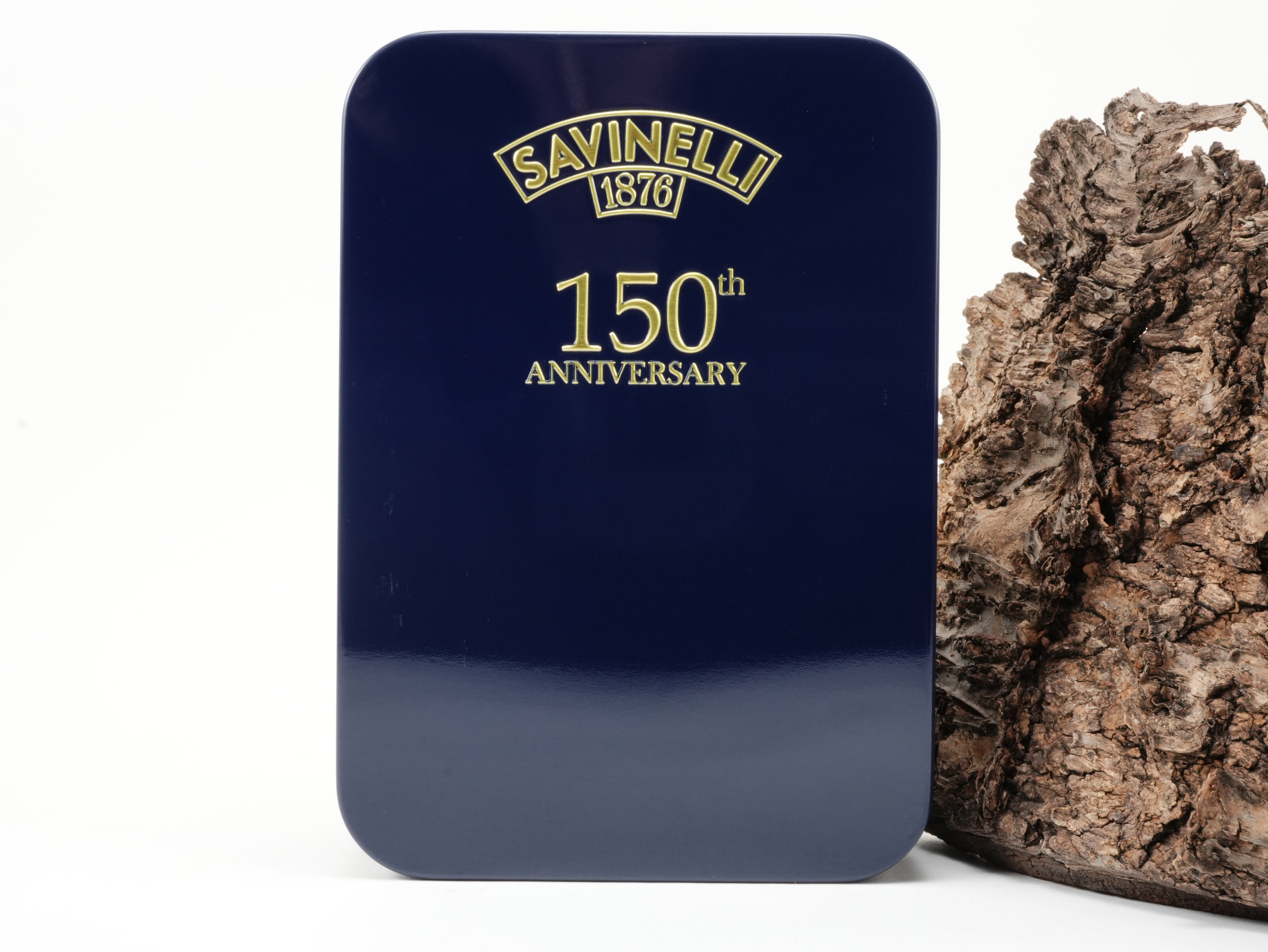Rechteckige tiefblaue Savinelli 150th Anniversary Tabakdose mit goldener Jubiläums-Prägung vor Baumrinde – Titelbild