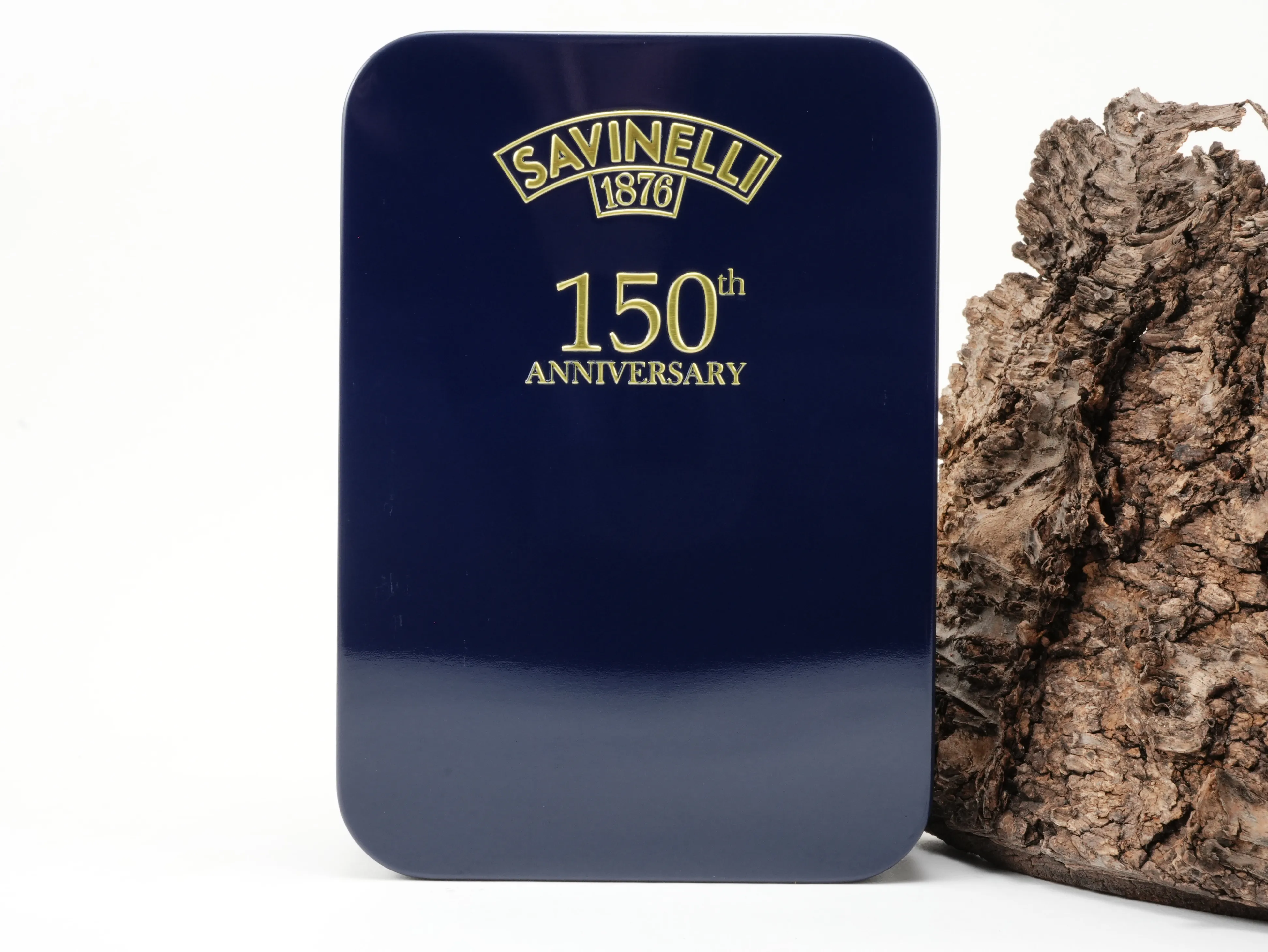 Rechteckige tiefblaue Savinelli 150th Anniversary Tabakdose mit goldener Jubiläums-Prägung vor Baumrinde – Titelbild